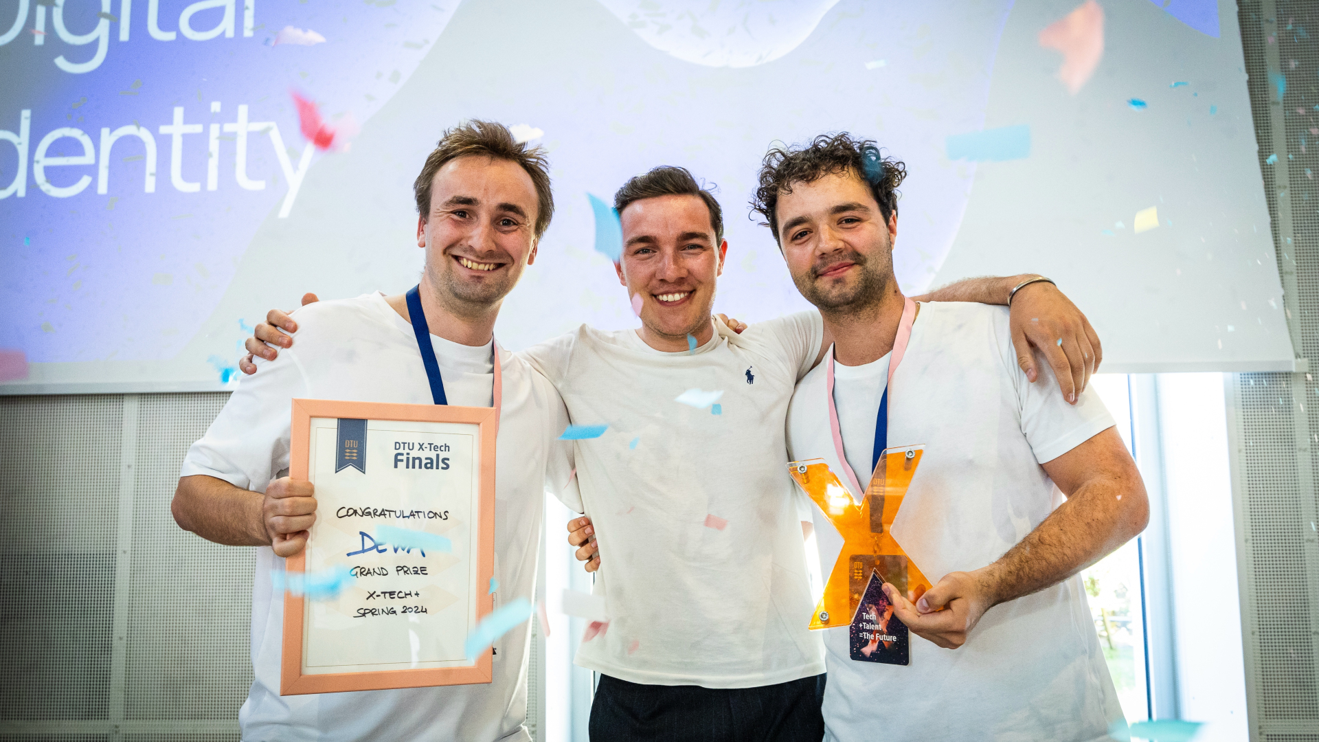 Tre unge mænd står side ved side mens konfetti regner ned over dem - den ene holder et stort orange X  og den anden et certifikat på at de har vundet X-Tech-finalen.