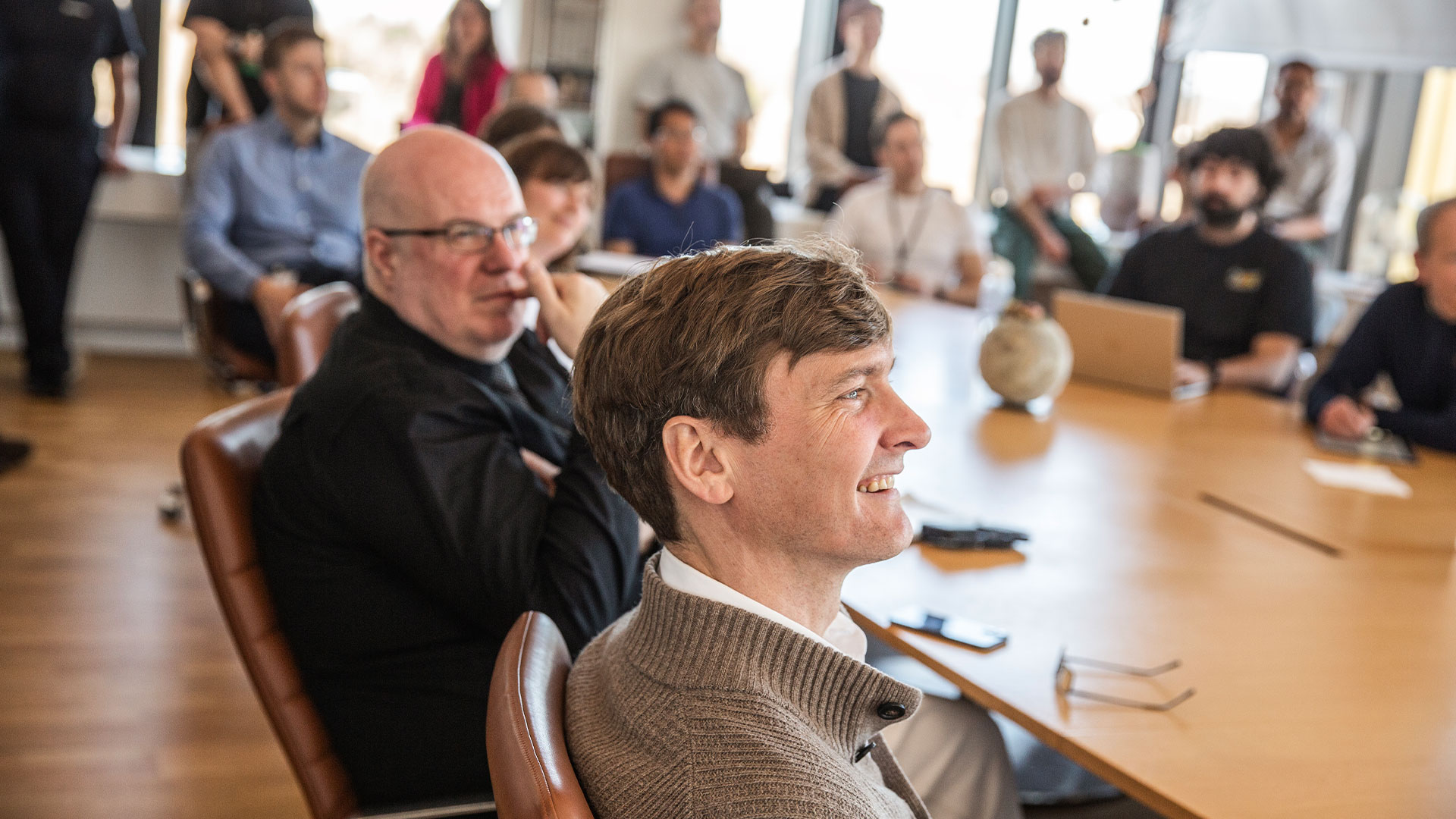 En mand med et bredt smil sidder blandt en gruppe mennesker ved et bord. Foto: Ditte Valente