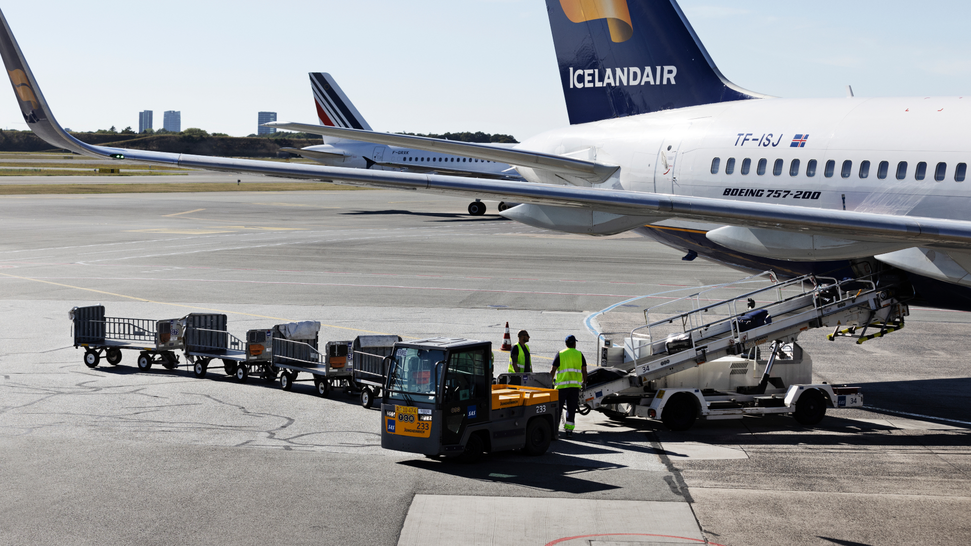 Bagenden af et passagerfly, hvor et transportbånd sender kufferter ud fraflyet og en bagagevogn holder og venter.