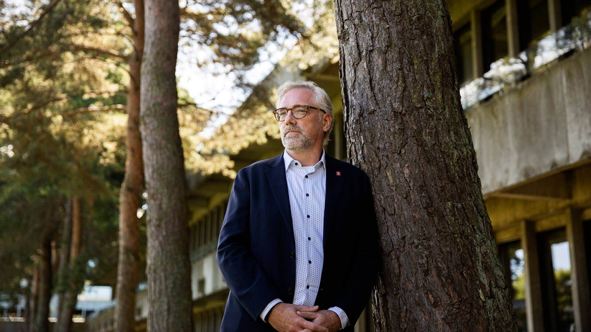Mand iført jakkesæt læner sig op ad træ, der står foran en betonbygning. Han bærer briller og kigger let til højre. 